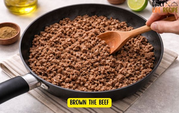 Ground beef browning in a skillet as it’s broken up with a spatula, cooking in olive oil on the stovetop for a burrito bowl filling.